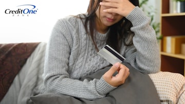 A young woman covers her face while sitting on her couch and holding a credit card in one hand, indicating that she may be confused or stressed about her credit.