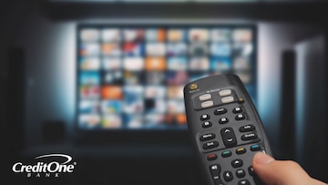 A man’s hand uses a remote to navigate various subscription services on a smart TV, which could impact his personal finances.