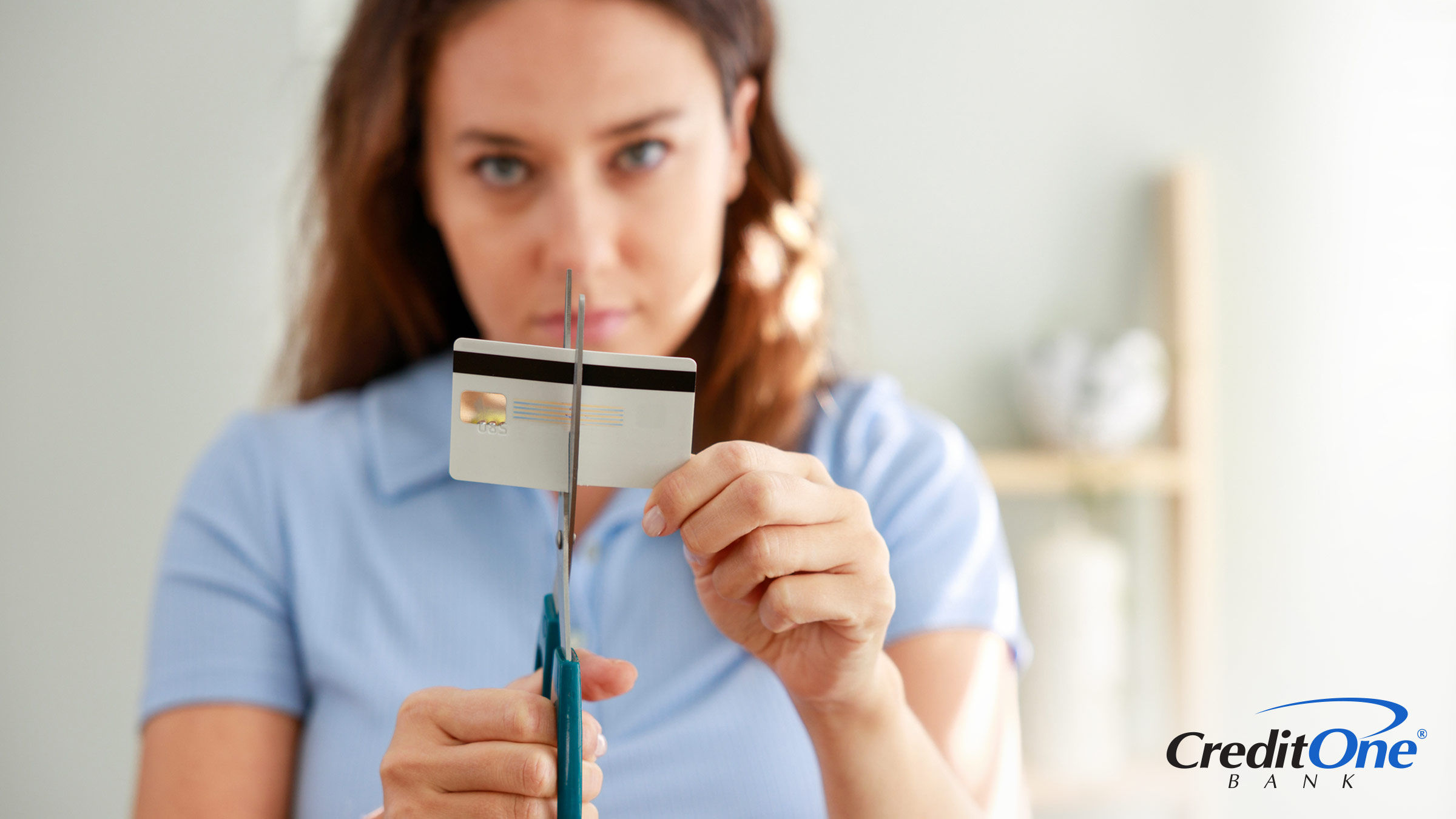 A young woman cuts up her credit card with a pained look on her face, likely wondering if closing the card will hurt her credit score.