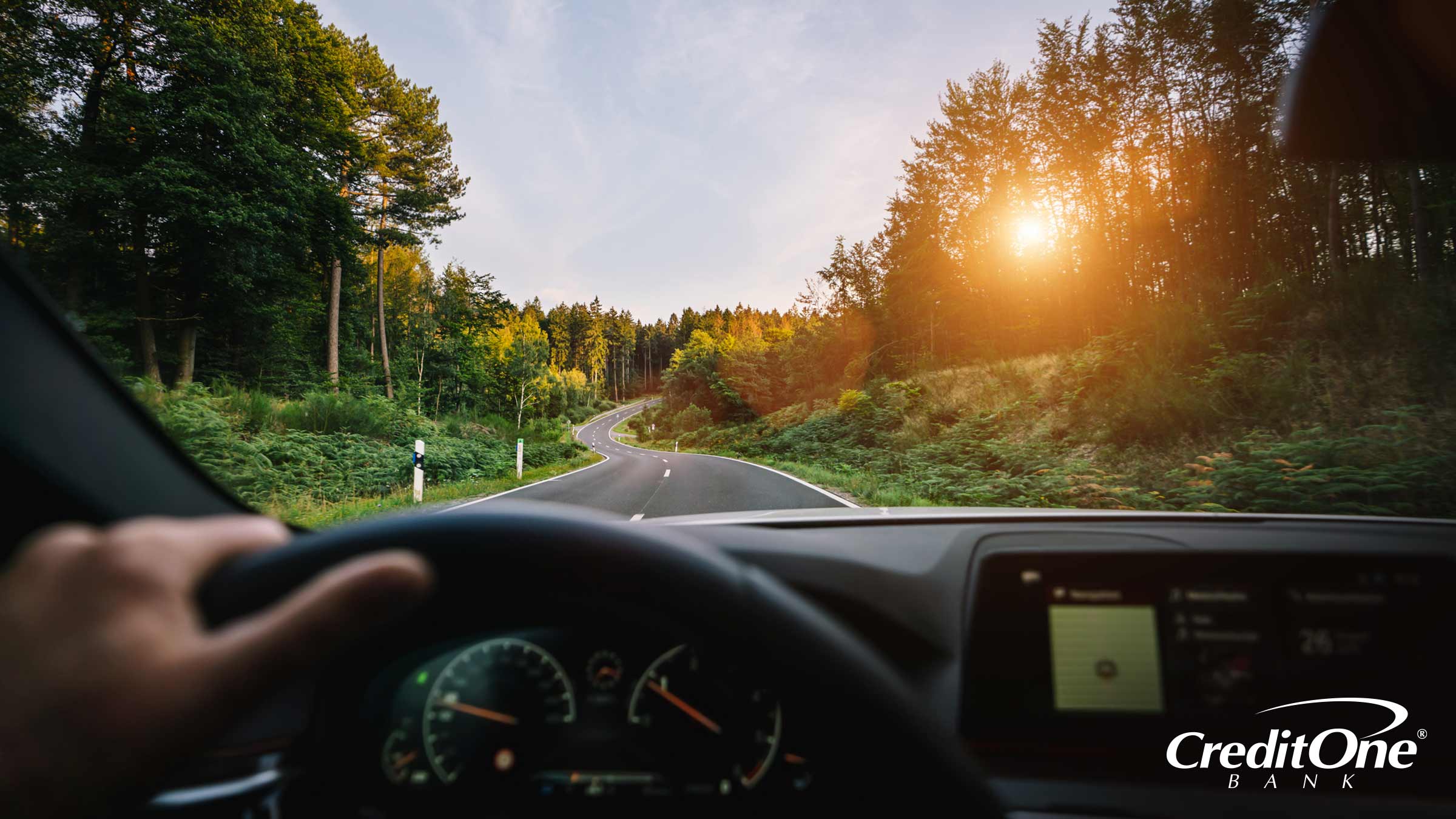 Someone behind the wheel, possibly a young Gen Z adult, is driving down a road with the sun slowly setting, guiding their way through the surrounding trees.