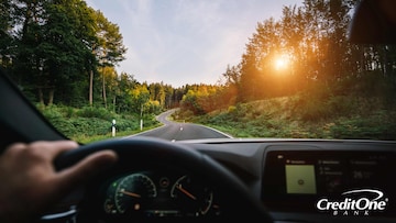 Someone behind the wheel, possibly a young Gen Z adult, is driving down a road with the sun slowly setting, guiding their way through the surrounding trees.