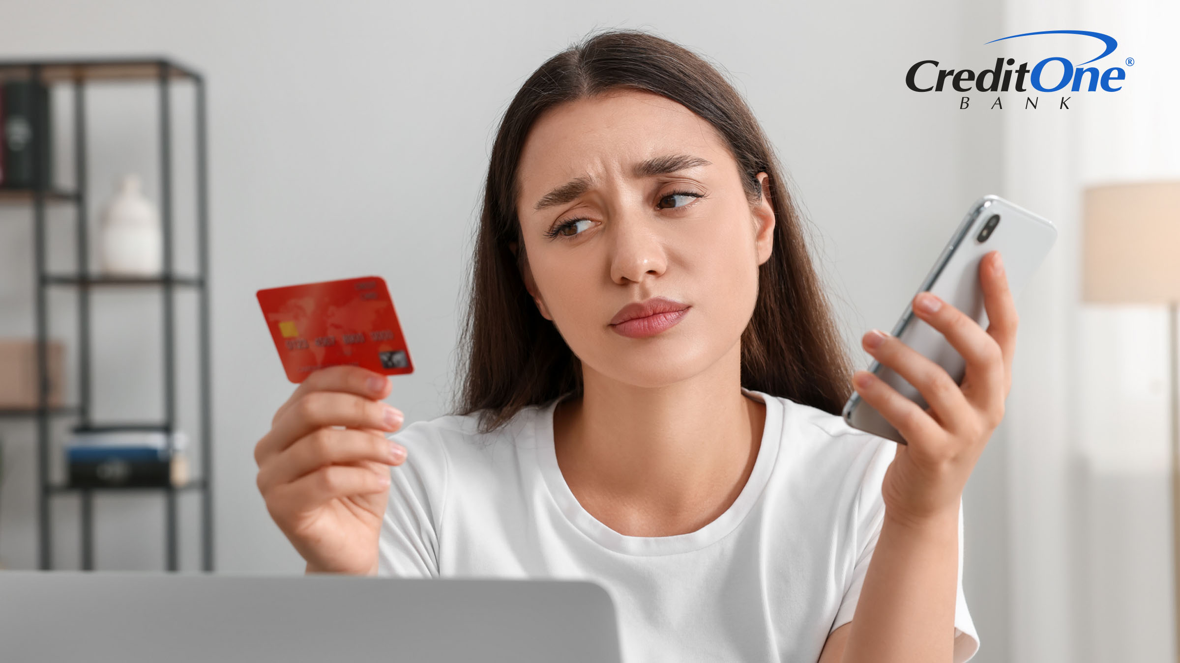 A young woman at a laptop scowls as she looks at her credit card in one hand while holding her mobile phone in the other, likely wondering whether her credit utilization ratio is too high.