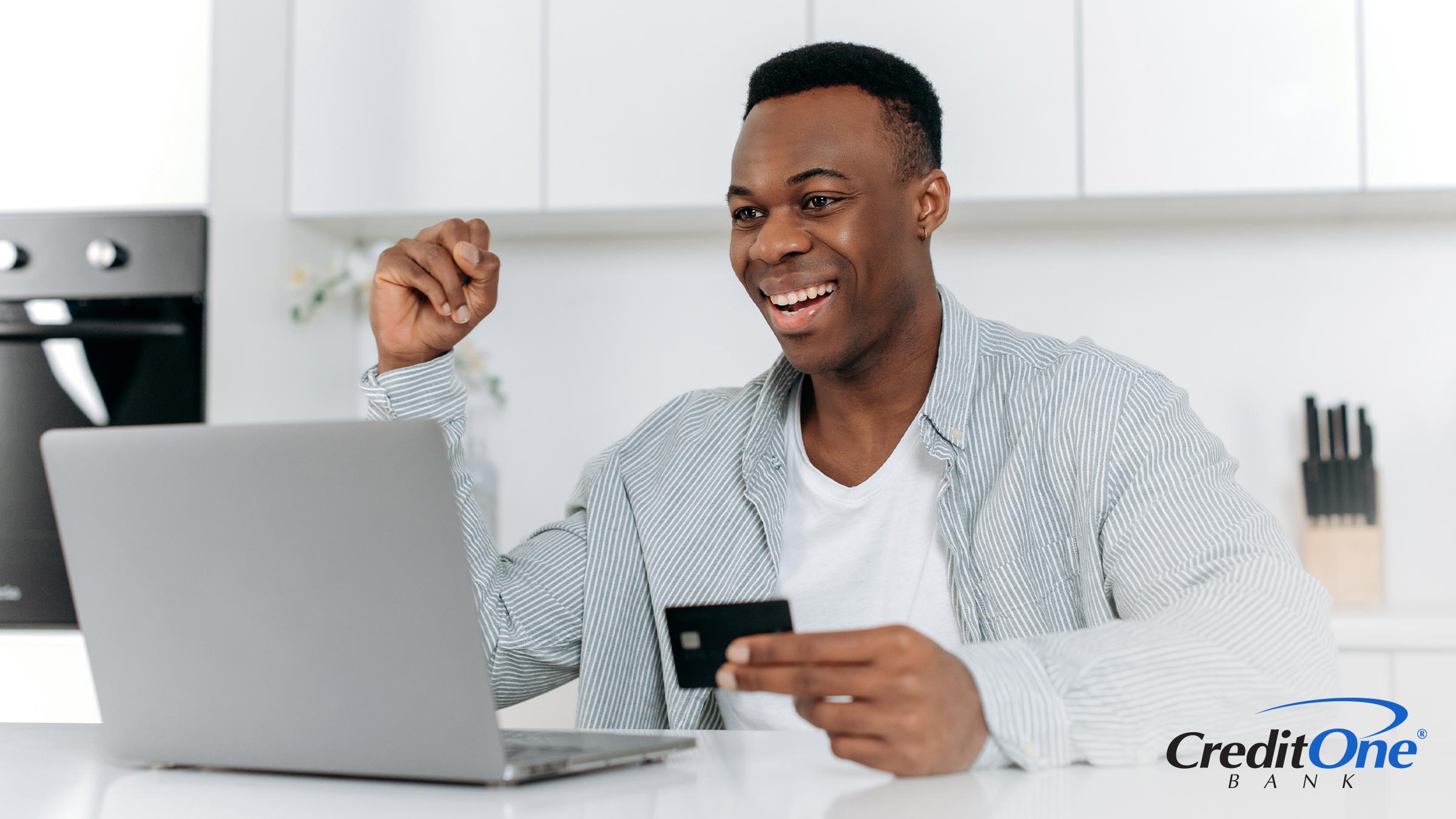 A young man smiles and pumps his fist in excitement as he holds a new credit card while using his laptop.