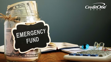 A cash-filled glass jar labeled “Emergency Fund” sits on a desk, representing one of the savings habits consumers might use to prepare for unforeseen expenses.