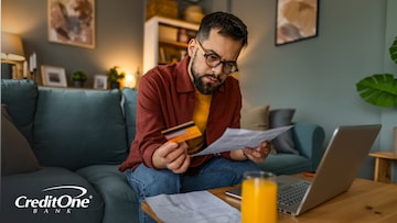 A man sits in his living room, holding up a credit card while reviewing his statement with a concerned expression. He may have noticed something unfamiliar, like a negative balance.