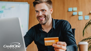 A young man smiles as he sits at his desk holding a credit card and using a laptop, which is a great tool for experiencing the benefits of online banking.