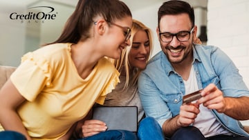 Two smiling young women sit on a couch beside a smiling young man who’s showing them a credit card like he’s choosing it as the right one.