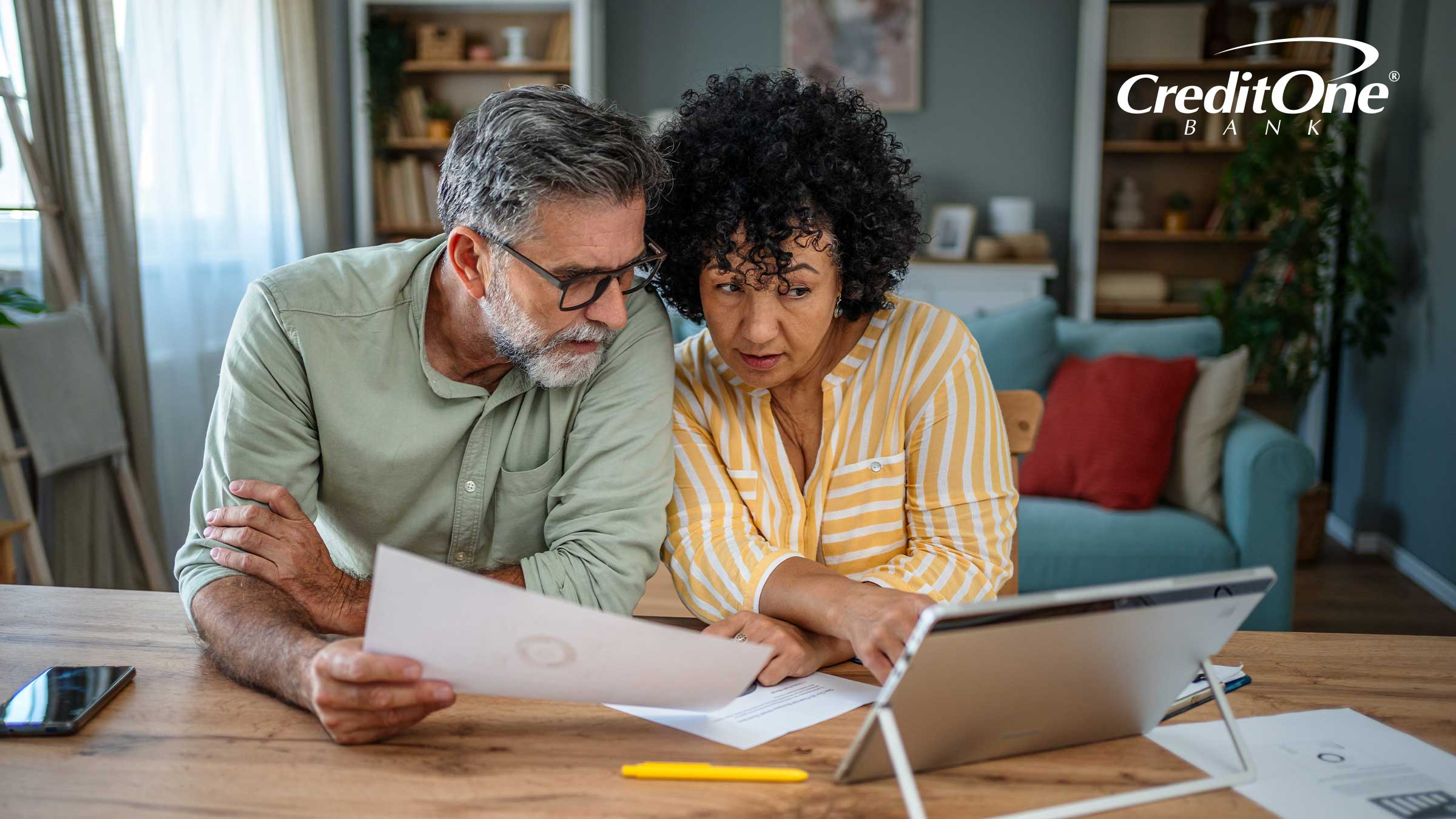 A mature couple sits at their kitchen table with tablet, phone, and financial documents. They may be evaluating savings account rates to determine if they’ll move some money or switch banks.