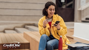 A smiling young woman sits on the steps with her backpack, laptop, and water bottle beside her. She holds a credit card in one hand and her phone in the other as if she’s monitoring her accounts while building credit.
